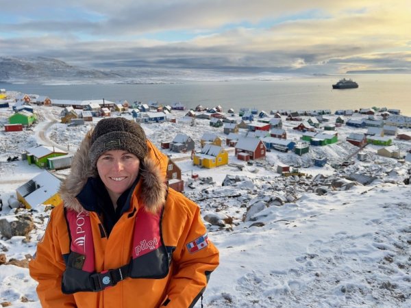 Tara in Greenland Woman in orange polar jacket, standing on a snowy hill in front of a village